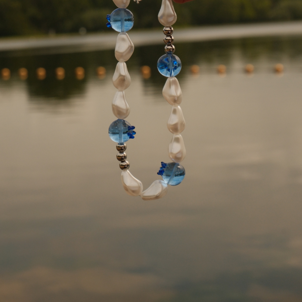 A beaded necklace with  irregular white baroque pearls, blue berrie shaped beads, and small silver spacer beads, photographed outdoors against a blurred lake background.