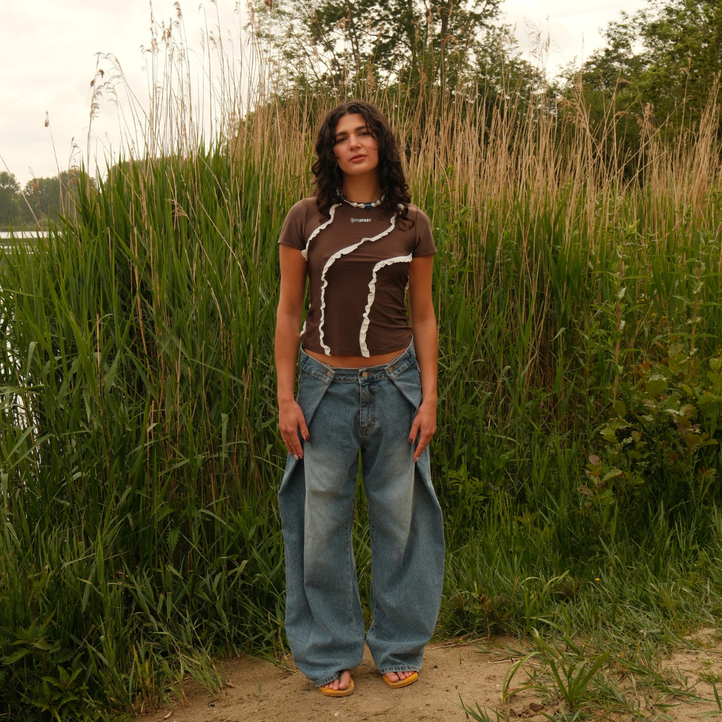 A woman stands outdoors in front of tall reeds wearing a brown short-sleeve crop top. She is also wearing a necklace. 